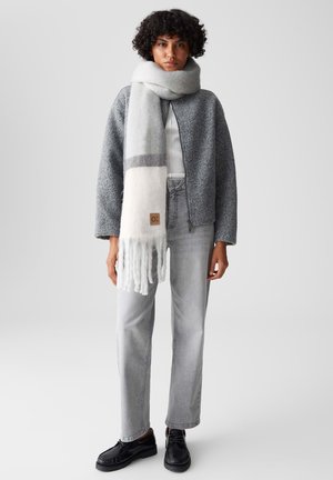 Grau gestrickte Jacke, weißer Schal mit grauem Streifen, graue hochtaillierte Jeans und schwarze Schuhe. Der Schal hat fransige Enden und ein Etikett.