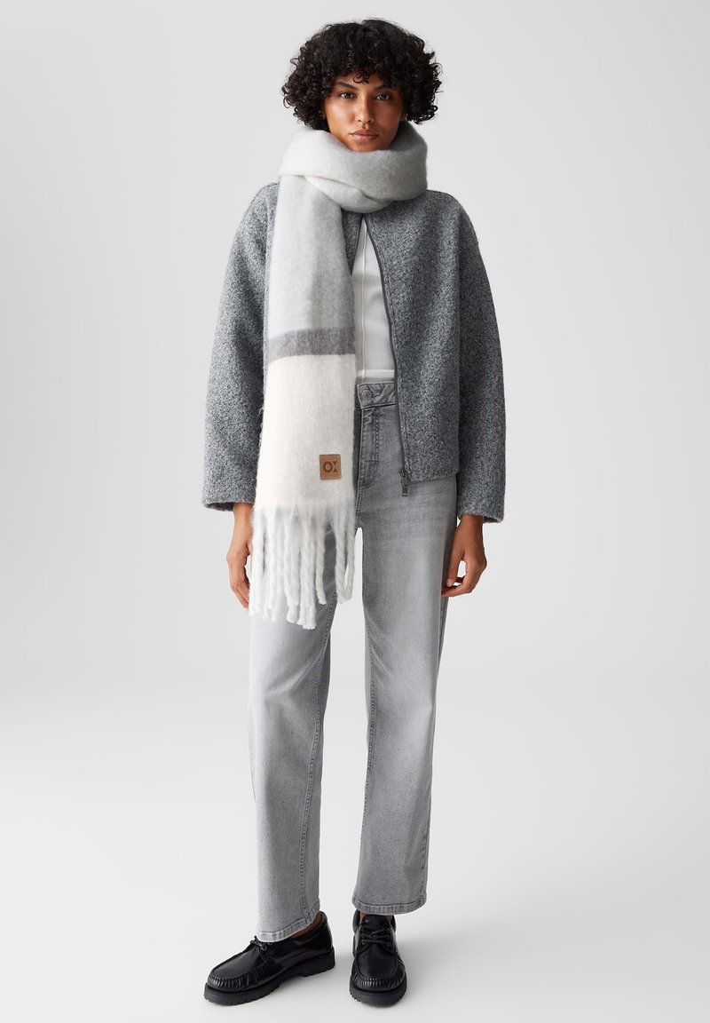 Grijze gebreide jas, witte sjaal met grijze streep, grijze high-waisted jeans en zwarte schoenen. De sjaal heeft franjes aan de uiteinden en een label.