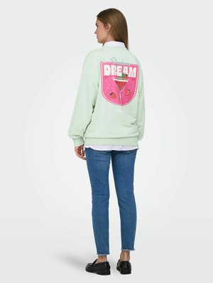 Vrouw draagt een lichtgroene trui met op de achterkant een roze "Strawberry Dream"-grafiek en fruitillustratie, blauwe spijkerbroek en zwarte schoenen.