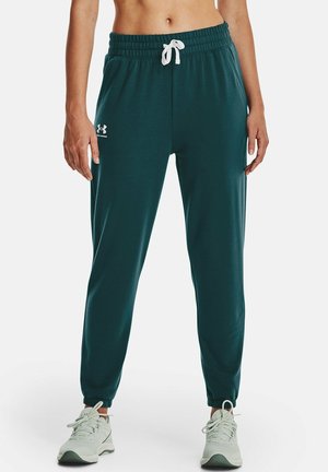 Žena nosící tyrkysové tepláky Under Armour s elastickým pasem a bílým šňůrkem, doplněné bílými sportovními botami.