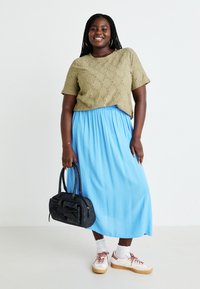 Green knitted short-sleeve top with diamond patterns, paired with a flowing blue maxi skirt, black handbag, and white sneakers with pink accents.