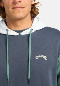 Granatowy hoodie z białym sznurkiem i zielonymi akcentami. Zawiera haft "BILLABONG" w kremowym kolorze. Materiał to teksturowana mieszanka bawełny.