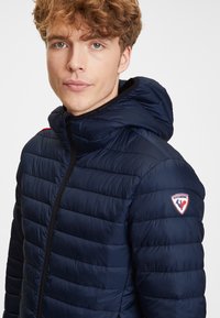 Marinblå pufferjacka med dragkedja, har en huva, horisontellt quiltad yta och ett logotypmärke på vänster axel.