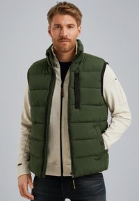 Green padded vest with a high collar, black accents, zippered chest pocket, and snap buttons. Worn over a textured beige sweatshirt.