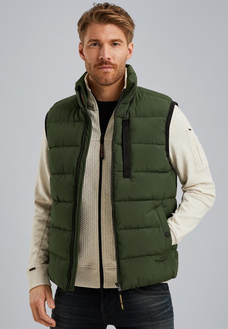Green padded vest with a high collar, black accents, zippered chest pocket, and snap buttons. Worn over a textured beige sweatshirt.