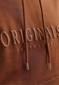Gros plan sur un sweat à capuche marron avec le texte en relief "ORIGINALS" et un texte plus petit "JACK & JONES", comprenant deux cordons avec des embouts argentés.