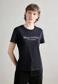 Juodas medvilnės marškinėlis su apvalia iškirpte ir trumpomis rankovėmis, priekinėje pusėje su baltais užrašais "Marc O'Polo EST. IN STOCKHOLM".