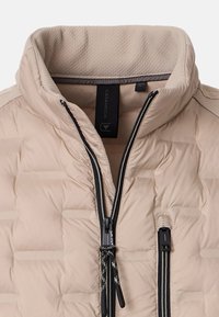 Beige pufferjacka med hög krage, med quiltad textur, svarta dragkedjor och diagonala sömmar på ärmarna.