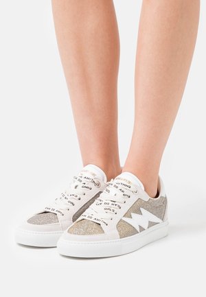 Benen met witte en glinsterende lage sneakers met witte bliksemschichtdetail en veters bedrukt met "Meisjes kunnen alles."