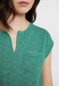 Vihreä raidallinen lyhythihainen paita, jossa on V-aukko, pieni rintatasku ja rento istuvuus. Valmistettu kevyestä puuvillamateriaalista.