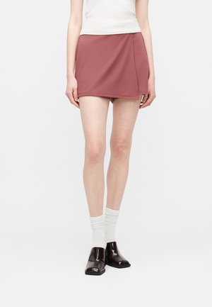 Persoon draagt een roosroze wikkelstijl minirok, witte sokken, donkerbruine schoenen met een vierkante neus en een witte mouwloze top tegen een effen achtergrond.