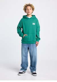 Sweat à capuche vert avec une poche avant et un patch logo, associé à un jean bleu clair ample et des baskets noires avec des accents blancs.