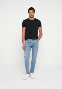 Czarny t-shirt z krótkim rękawem w połączeniu ze światło niebieskimi prostymi dżinsami i białymi tenisówkami, na tle jednolitej białej ściany.