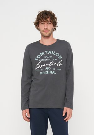 Hombre con cabello rizado que lleva una camisa de manga larga gris oscuro con el texto "Tom Tailor Essentials Original", de pie frente a un fondo blanco liso.