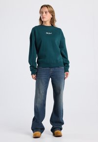 Sudadera verde bosque con puños y dobladillo acanalados, que presenta texto bordado en blanco. Combinada con jeans de pierna ancha azul claro y calzado beige.