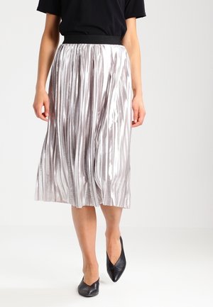 Woman wearing a shiny silver pleated midi skirt, black top, and black pointed-toe flats, walking against a plain white background.