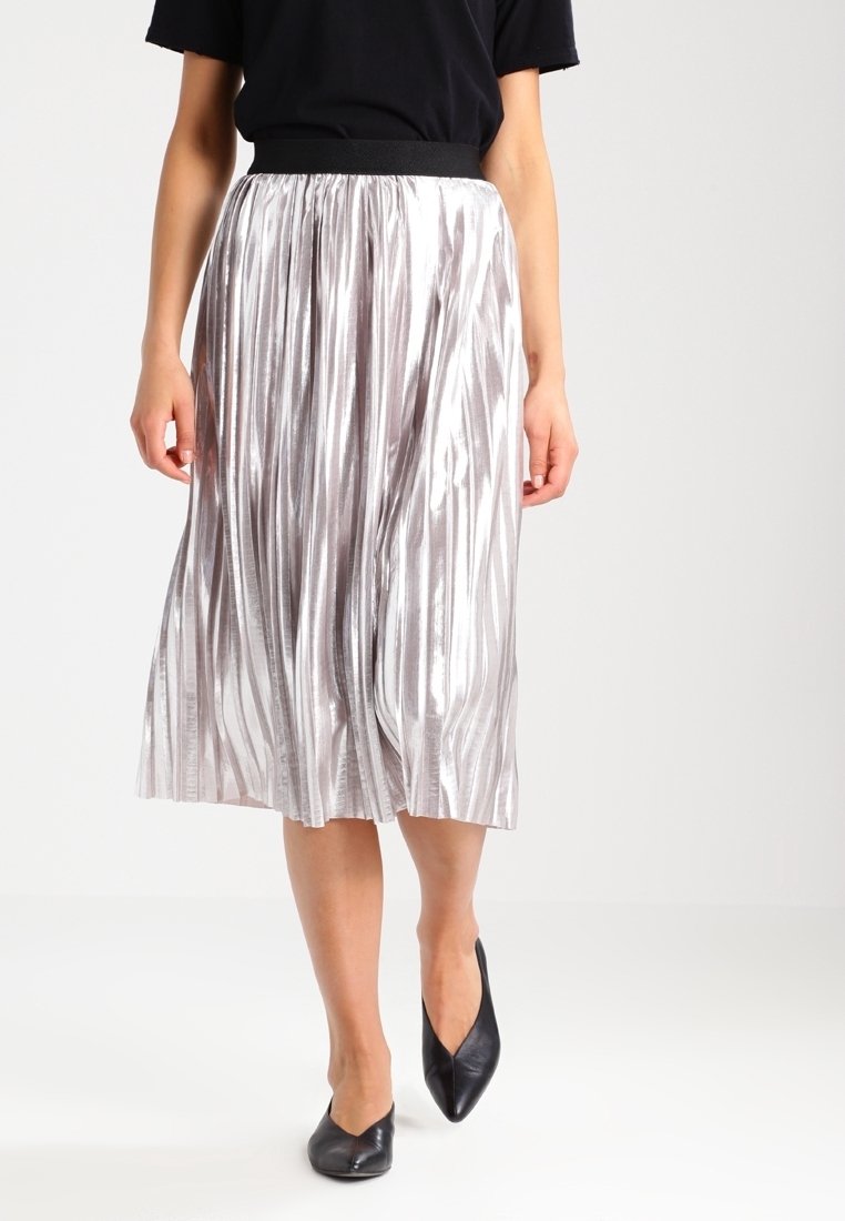 Femme portant une jupe midi plissée argentée brillante, un haut noir et des ballerines noires à bout pointu, marchant sur un fond blanc uni.