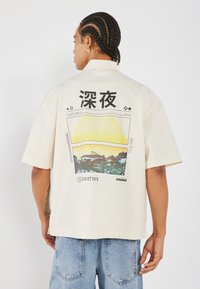 Cămașă cu mânecă scurtă, de culoare crem, care prezintă un imprimeu grafic colorat pe spate, cu text în limba japoneză și elemente de design peisagistic.