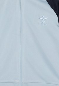 Lichtblauwe ritsjack gemaakt van zachte stof, met een donkerblauwe accent op één schouder en een geborduurd logo op de borst.