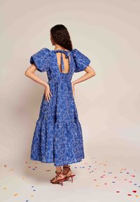 Μάξι φόρεμα με μπλε floral σχέδιο, φουσκωτά μανίκια, ανοιχτή πλάτη και κορδόνι κλεισίματος. Υφή υφάσματος και στρωτή φούστα. Κόκκινες ψηλοτάκουνες στιλέτο.