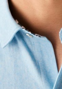 Camisa azul claro con un patrón de rayas, que presenta un cuello abotonado y un ribete interior con un diseño contrastante. Textura suave.