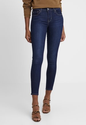 Mørkeblå jeans med tætsiddende pasform, båret med brune sandaler med leopardprint og hæl med stropper på en lys baggrund.