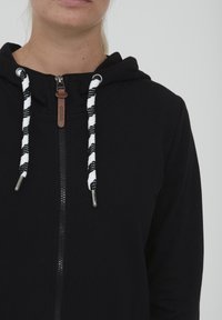 Juodas zip-up didelis su gobtuvu, su juodai baltais dryžuotais virvutėmis ir odiniu akcentu ant užtrauktuko.