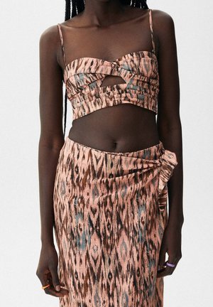 Woman wearing a sleeveless cropped top and matching wrap skirt with geometric pink, brown, and blue pattern, standing against a plain background.