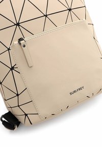 Sac à dos beige avec des lignes géométriques noires et une poche avant zippée étiquetée « SURI FREY ».