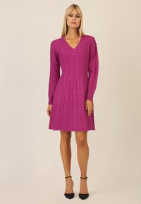 Fuchsia gestricktes Kleid mit langen Ärmeln, V-Ausschnitt und Zopfmuster. Der schwingende Rock und die gerippten Bündchen verleihen dem Design eine besondere Note. Schwarze spitze Absätze.