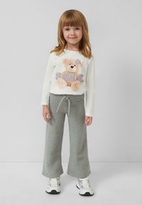Weißes Langarmhemd mit einem Teddybär-Graphic und dem Wort "LÄCHELN." Graue weit geschnittene Fleece-Hose. Sneakers mit weißen und schwarzen Akzenten.