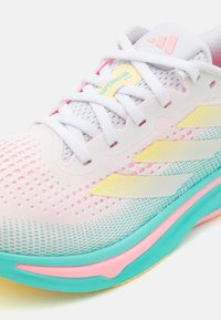 Primer plano de una zapatilla de correr Adidas Supernova Rise blanca con detalles en rosa pastel, amarillo y verde claro, y cordones blancos sobre un fondo blanco.