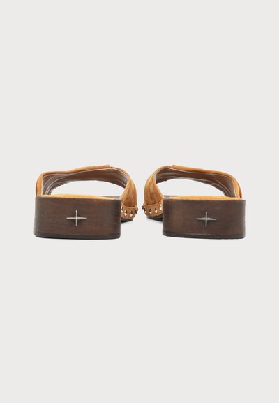 Paire de chaussures en daim marron à enfiler avec côtés cloutés et petits talons blocs en bois, vues de dos sur fond blanc.