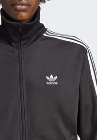 Detailní záběr na osobu v černé mikině Adidas se zipem, která má bílý trojitý pruhový design na ramenou a logo Adidas na hrudi.