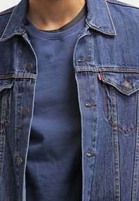 Giacca di jeans con bottoni metallici, colore blu, e colletto a punta, sovrapposta a una maglietta blu navy a girocollo in un tessuto morbido.