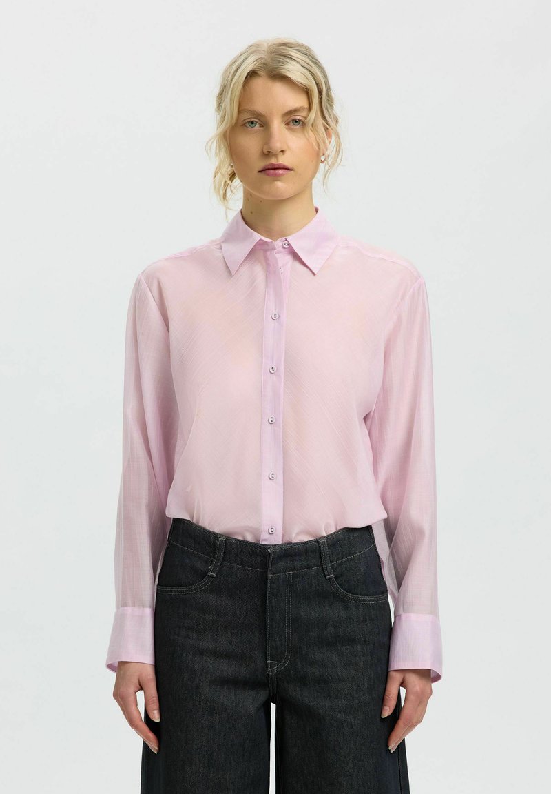 Femme aux cheveux blonds portant une chemise rose clair à manches longues boutonnée, rentrée dans un jean sombre taille haute, debout devant un fond uni.