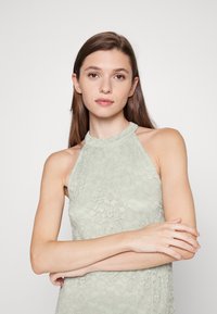 Vestido halter verde claro em renda com um padrão floral texturizado, apresentando um decote alto e um design de ombros recortados.