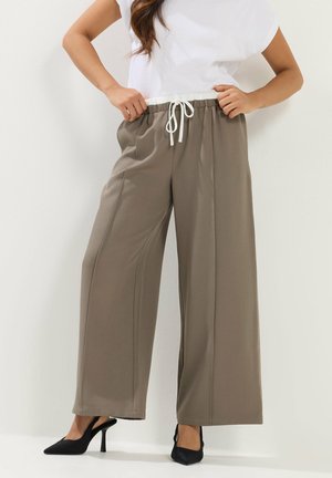 Vrouw draagt taupekleurige wijde broek met witte trekkoorden en zwarte hooggehakte slingback schoenen, staand tegen een effen muur.