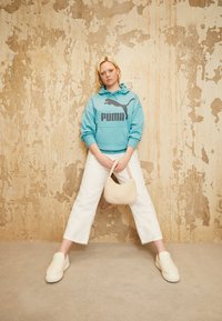 Svetlo modrá mikina s kapucňou a čiernym logom Puma, biele široké nohavice, krémová chlpatá taška a zodpovedajúce slip-on topánky na textúrovanom pozadí.