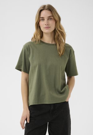 Femme aux cheveux châtain clair portant une chemise à manches courtes vert olive et un pantalon noir, debout devant un fond clair uni.