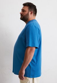 Blauwe T-shirt met korte mouwen, gemaakt van zachte katoen, met een ontspannen pasvorm en een ronde halslijn, gedragen met lichtgekleurde broeken.