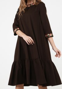 Kvinna i en lös mörkbrun klänning med volang nederkant och blommig broderi vid halsringning och ärmar, poserar mot en vit bakgrund.