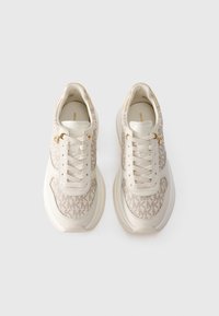 O pereche de adidași crem și bej cu un model monogram și accente aurii pe orificiile pentru șireturi, văzuți dintr-o parte de sus pe un fundal alb.