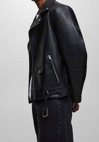 Chaqueta de motociclista de cuero negro con cremalleras, bolsillos laterales y cintura con cinturón. Presenta un diseño clásico con un ajuste ligeramente holgado.