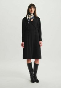Robe enveloppante noire à manches longues et taille cintrée, dotée de boutons croisés. Associée à des bottes noires jusqu'aux genoux et à une écharpe à motifs.
