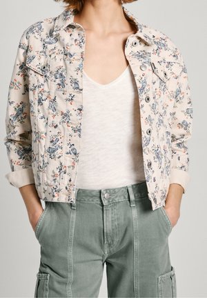 Žena má na sobě lehkou květovanou zapínanou bundu přes bílou tričko s výstřihem do V a zelené cargo kalhoty s rukama v kapsách.