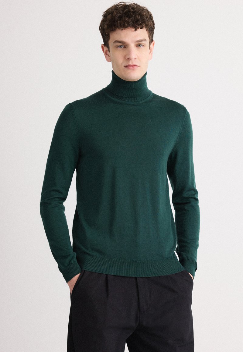 Dunkelgrüner Rollkragenpullover aus einem glatten Strickstoff, mit langen Ärmeln und taillierter Silhouette. Kombiniert mit schwarzen Hosen.