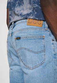 Vista trasera de una persona que lleva puestos unos jeans de Lee de color azul claro y una camiseta de tie-dye azul, centrando la atención en el bolsillo trasero de los jeans y la etiqueta de la marca.