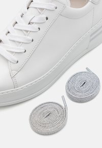 Witte leren sneaker met platte witte veters en zilverkleurige accenten. Twee zilveren veters liggen naast de sneaker.