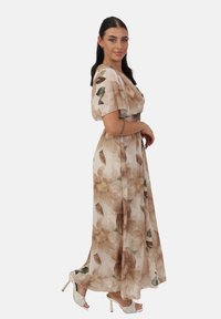 Robe maxi en chiffon beige clair, ornée de grands motifs floraux marron, avec des manches courtes et flottantes, une taille cintrée et des fentes sur les côtés. Avec des talons blancs.